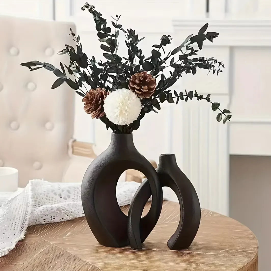 Modern black ceramic vase with abstract cutouts, holding dried eucalyptus, pinecones, and white bloom on wooden table